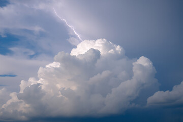 Dramatic sky view featuring a bright lightning bolt illuminating a large, fluffy white cloud formation amidst darker storm clouds.