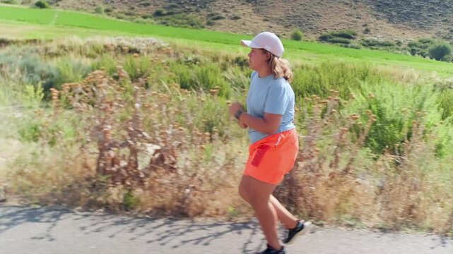Side view. A overweight girl runs along a road through open terrain at the base of the mountains. Regular training helps her lose weight, overcome barriers, and work toward better health and new goals