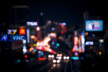 Blurred city lights create a vibrant bokeh effect in the night cityscape. Car headlights and signs illuminate the scene with streaks of color against the dark sky.