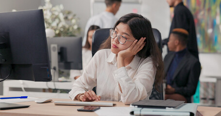 A tired office secretary in a white shirt sits at her desk, looking at the computer monitor. She supports her face with her hand, feeling bored and longing for the workday to be over.