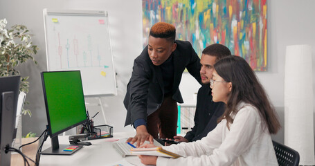 Fototapeta premium A male manager in a suit leads training, engaging employees with questions. He assists them in developing a project strategy, pointing out important details on the computer monitor and explaining the