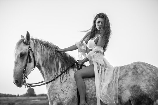 Beautiful brunette woman with long hair ,white bikini and white shirt rides  on a white horse in the river. Sensual and attractive girl  interacting and ride a horse in the water at sunset