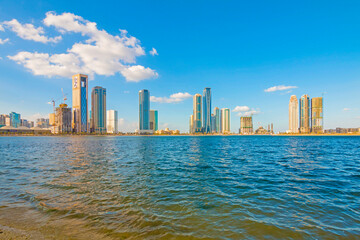 Sharjah city skyline at sunrise with golden light reflecting on modern skyscrapers. Serene beach with gentle waves. Peaceful morning atmosphere in the UAE’s cultural capital.