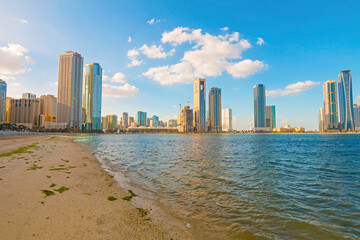 Sharjah city skyline at sunrise with golden light reflecting on modern skyscrapers. Serene beach with gentle waves. Peaceful morning atmosphere in the UAE&rsquo;s cultural capital.