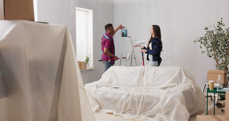 A couple in love stands with paint rollers in front of furniture covered with protective film. They are preparing to paint the walls during a home renovation.