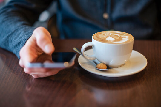 Young man holding a mobile phone with a cup of coffee on a table - Powered by Adobe