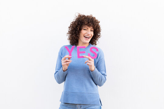 Woman with curly hair holding the word YES in front of a white wall, smiling and looking at the camera