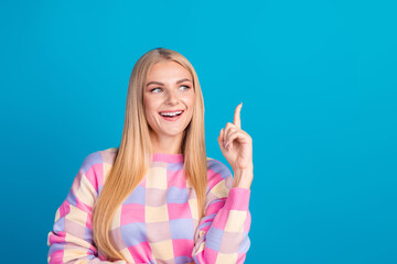 Smiling young woman pointing upward with a cheerful and charming expression on a bright blue...