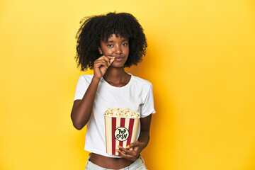 Teen girl with popcorn, cinema concept on yellow backdrop with fingers on lips keeping a secret. © Asier