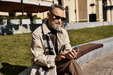 Obraz premium A senior man dressed in a chic outfit sits on a modern bench, interacts with his smartphone.