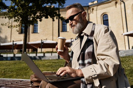 A dapper senior man sits casually on a bench, savoring his coffee while typing on his laptop.