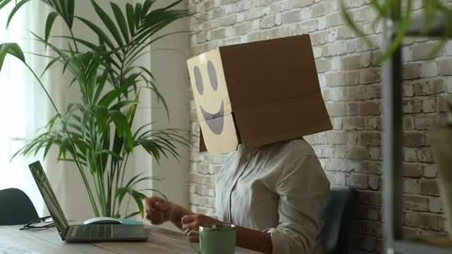 Corporate worker with hidden identity wearing a cardboard box on head while working at laptop in anonymous office space, symbolizing isolation, anonymity, digital privacy, and workplace alienation