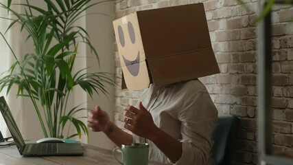 Office employee concealing identity with a cardboard box over head while typing on laptop at desk, representing faceless corporate work, digital isolation, and anonymity in modern business