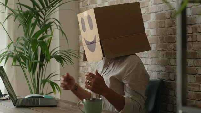 Office employee concealing identity with a cardboard box over head while typing on laptop at desk, representing faceless corporate work, digital isolation, and anonymity in modern business