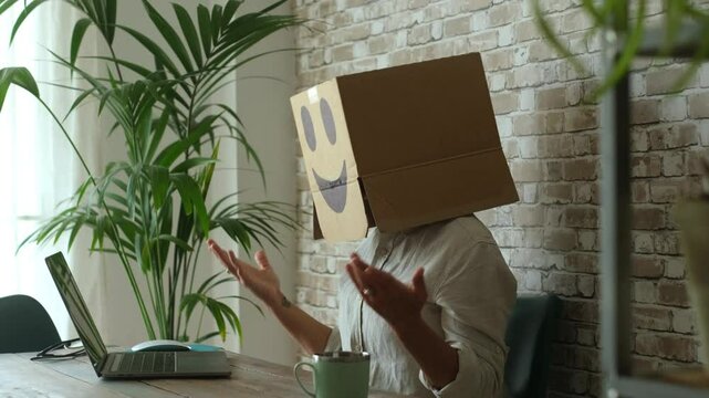 Professional in anonymous workplace using laptop while hiding identity with a cardboard box, reflecting themes of workplace alienation, surveillance, bureaucracy, and faceless employment