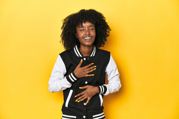 Teen girl in baseball jacket, yellow studio background laughs happily and has fun keeping hands on...