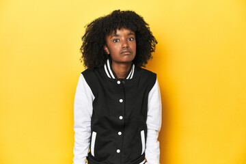 Teen girl in baseball jacket, yellow studio background shrugs shoulders and open eyes confused.