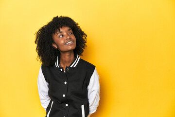 Teen girl in baseball jacket, yellow studio background relaxed and happy laughing, neck stretched...