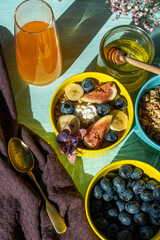 Granola and oatmeal with figs, blueberries and banana in a yellow bowl on a blue background are decorated with flowers. The concept of a healthy breakfast.