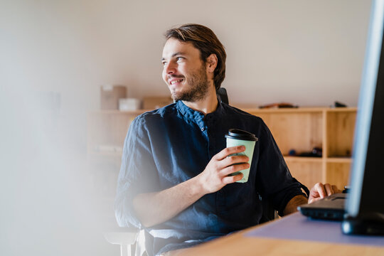 Smiling businessman with takeaway drink sitting at desk in wooden open-plan office