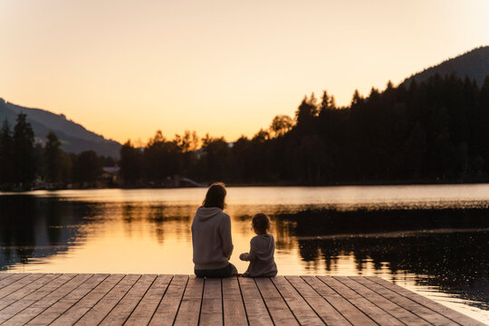 Mother and little daughter sitting together at end of lakeshore jetty at dusk