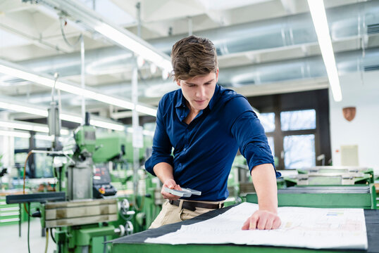 Male engineer examining blueprint while holding machine part in in industry
