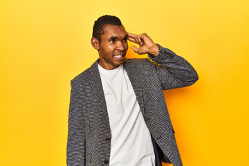 African American man in casual blazer, yellow studio, pointing temple with finger, thinking, focused on a task.