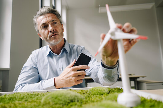 Businessman looking at wind turbine toy over moss frame while sitting at office