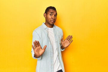 African American man in blue shirt, yellow studio, rejecting someone showing a gesture of disgust.