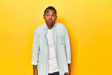 African American man in blue shirt, yellow studio, shrugs shoulders and open eyes confused.