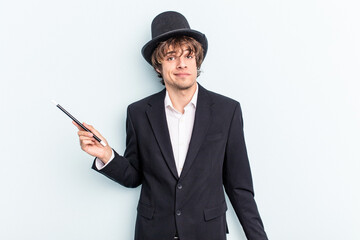 Young magician man holding magic cards isolated on blue background shrugs shoulders and open eyes...