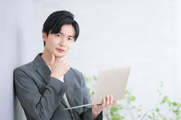 壁に寄りかかり立ってパソコンを持つカジュアルな若い男性の考えるビジネスマンのクローズアップのカメラ目線、 困る悩む表情