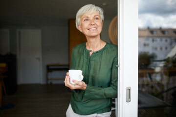 Senior woman standing with coffee cup by window at home