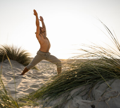 Shirtless young man practicing warrior position yoga at beach against clear sky during sunset