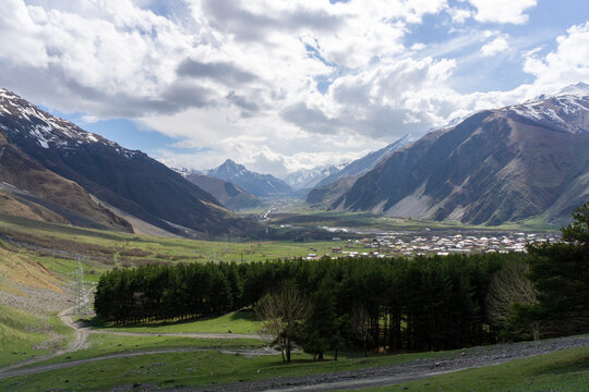 Roofs of houses in a gorge between mountains. A strip of coniferous trees in the foreground. High mountains and rocks covered with snow. Darial Gorge