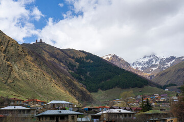 Mount Kazbek covered with snow and partly covered by clouds. Roofs of houses in Gergeti village and Stepantsminda, Holy Trinity Church on the hill