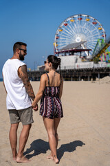 Tourist couple in love holding hands walking along the beach by the pier in Santa Monica, California.