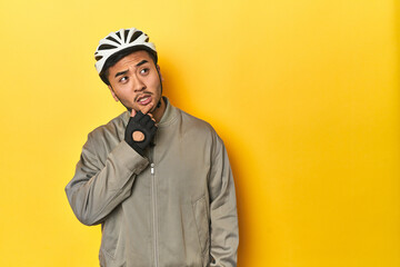 Asian man in bike helmet and gloves, yellow studio backdrop relaxed thinking about something...