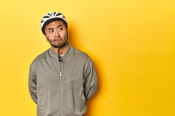 Asian man in bike helmet and gloves, yellow studio backdrop confused, feels doubtful and unsure.