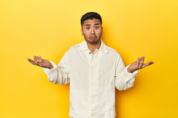 Asian man in crisp white shirt on yellow studio doubting and shrugging shoulders in questioning...