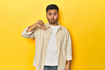 Casual Asian man with open shirt, white tee on yellow studio showing thumb down, disappointment...