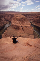 Unrecognizable young male tourist, sitting on the Horseshoe Bend while raising his arms in the U.S.