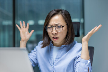 An Asian woman stares in disbelief at her computer screen in the office, experiencing frustration with an error or unexpected outcome.