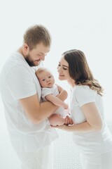 A happy young family mom dad and a small child smiling and kissing on a white isolated window background, a loving family threesome at home