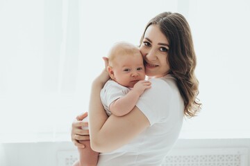 A happy young mother and a newborn baby smile, hug and kiss on a white isolated window background, a loving family at home