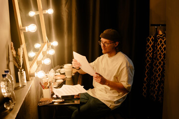Person reading script while seated beside an illuminated vanity mirror inside dressing room, creating a focused ambiance with a touch of anticipation