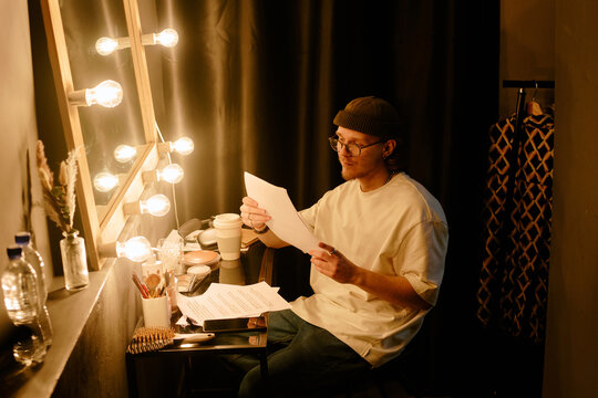 Person reading script while seated beside an illuminated vanity mirror inside dressing room, creating a focused ambiance with a touch of anticipation