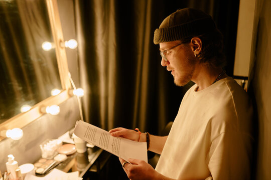Young male actor reading script in dressing room backstage before performance under warm light next to mirror lined with bulbs and various makeup products