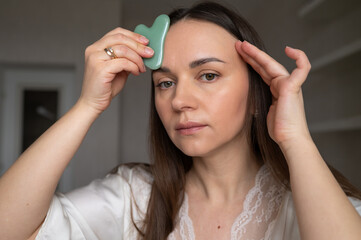 Fototapeta premium Young woman in silk robe doing facial massage with gua sha scraper while looking in mirror. Home skin care and aesthetic procedures