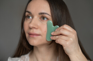 Young woman using a green gua sha scraper for facial massage, improving skin circulation and...